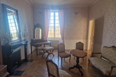 Salon ancien avec parquet en bois, mobilier classique, cheminée, miroir, coiffeuse et grande fenêtre avec rideaux roses.