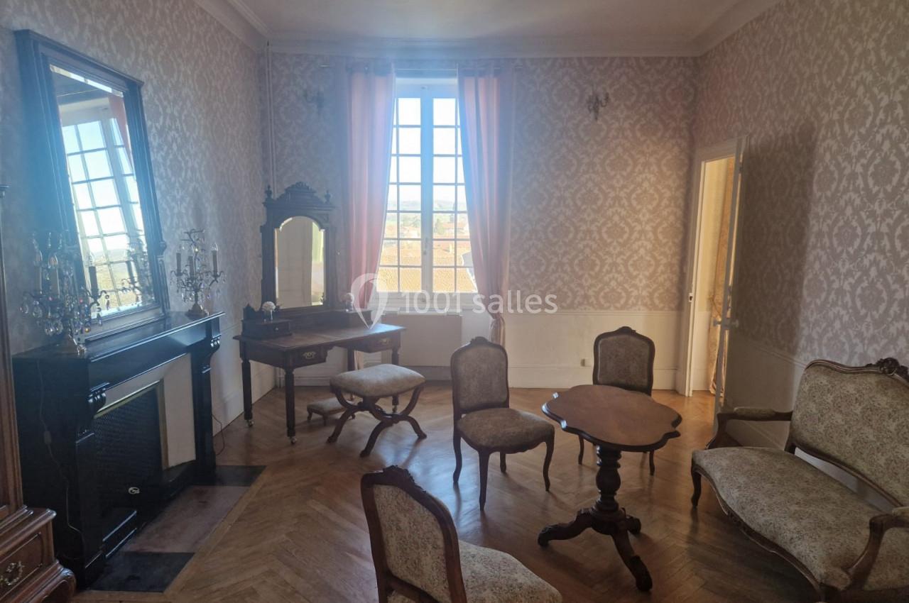 Salon ancien avec parquet en bois, mobilier classique, cheminée, miroir, coiffeuse et grande fenêtre avec rideaux roses.