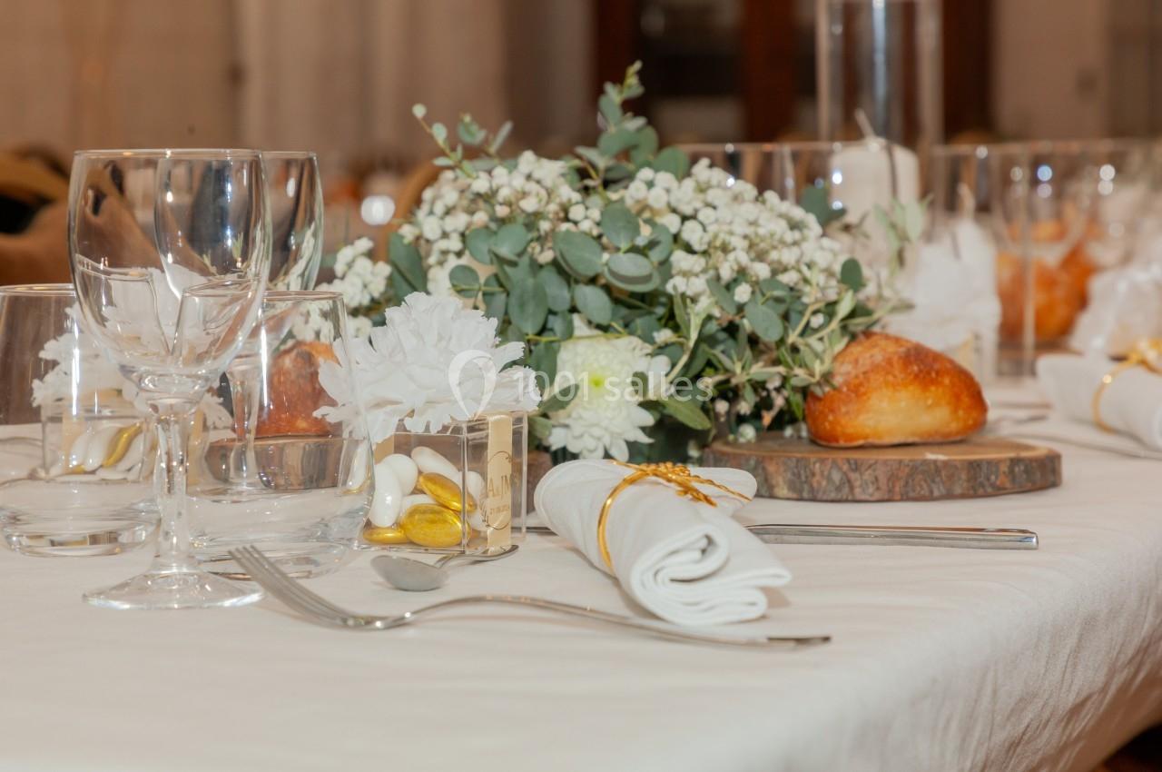 Table dressée avec des verres, couverts, serviettes pliées, fleurs blanches et verdure, accompagnée de pain et dragées.