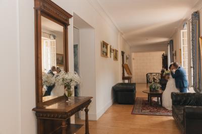 Salon ancien avec parquet en bois, mobilier classique, cheminée, miroir, coiffeuse et grande fenêtre avec rideaux roses.