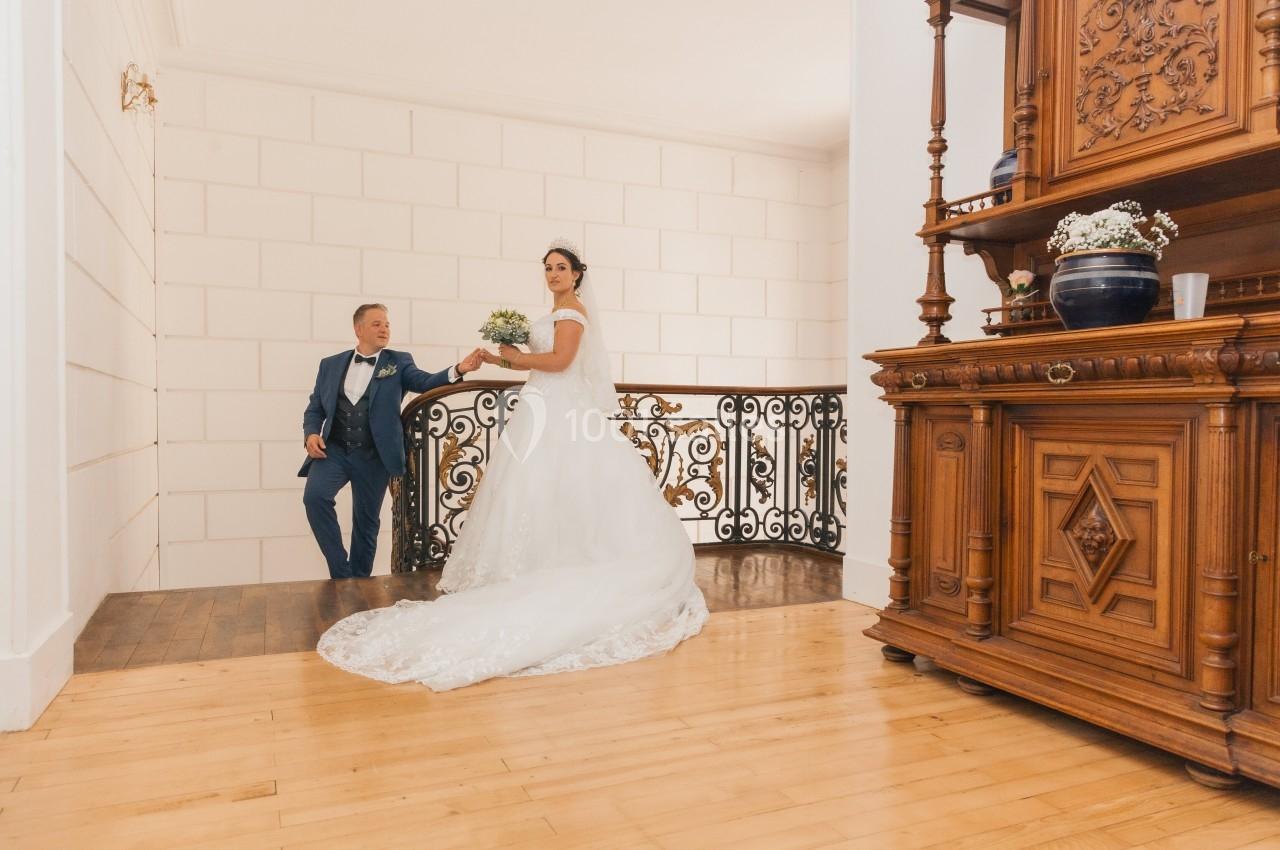 Un couple de mariés pose sur un escalier intérieur avec une rampe en fer forgé et un meuble en bois sculpté.