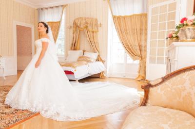 Salon ancien avec parquet en bois, mobilier classique, cheminée, miroir, coiffeuse et grande fenêtre avec rideaux roses.