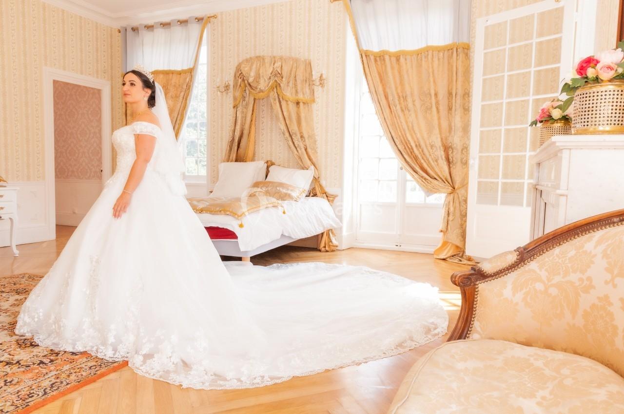 Femme en robe de mariée blanche dans une chambre lumineuse avec lit à baldaquin et rideaux dorés.
