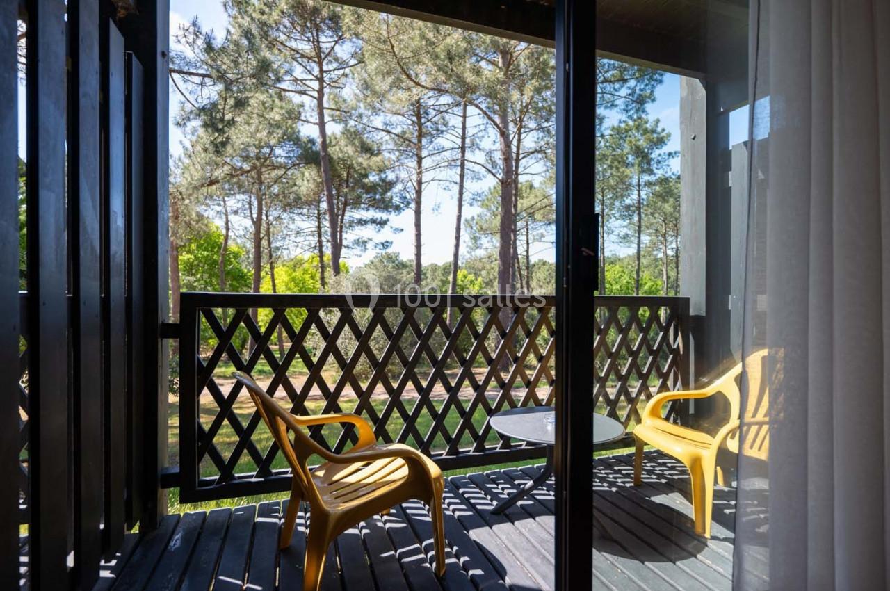 Vue depuis une porte-fenêtre sur une terrasse en bois avec deux chaises jaunes, une table et des arbres en arrière-plan.