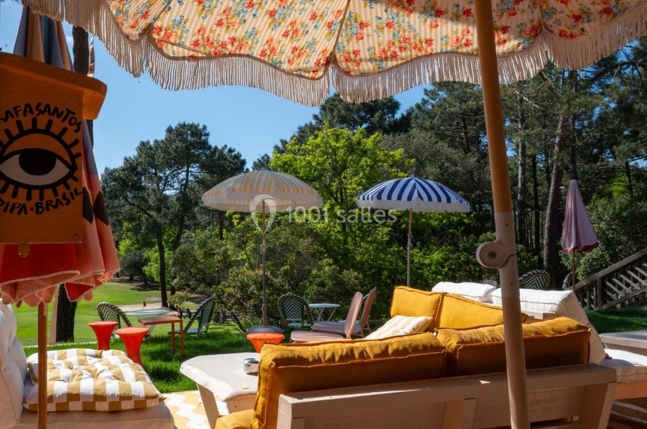 Terrasse ensoleillée avec canapés jaunes, parasols colorés et vue sur un jardin verdoyant entouré d'arbres.