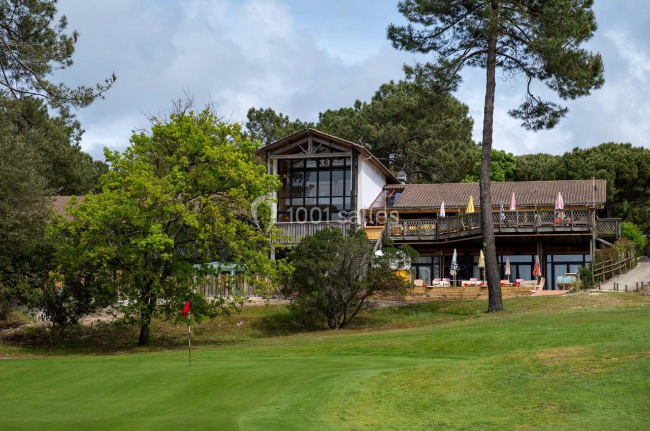 Bâtiment en bois avec terrasse entouré de pins, situé près d'un terrain de golf avec un drapeau rouge.