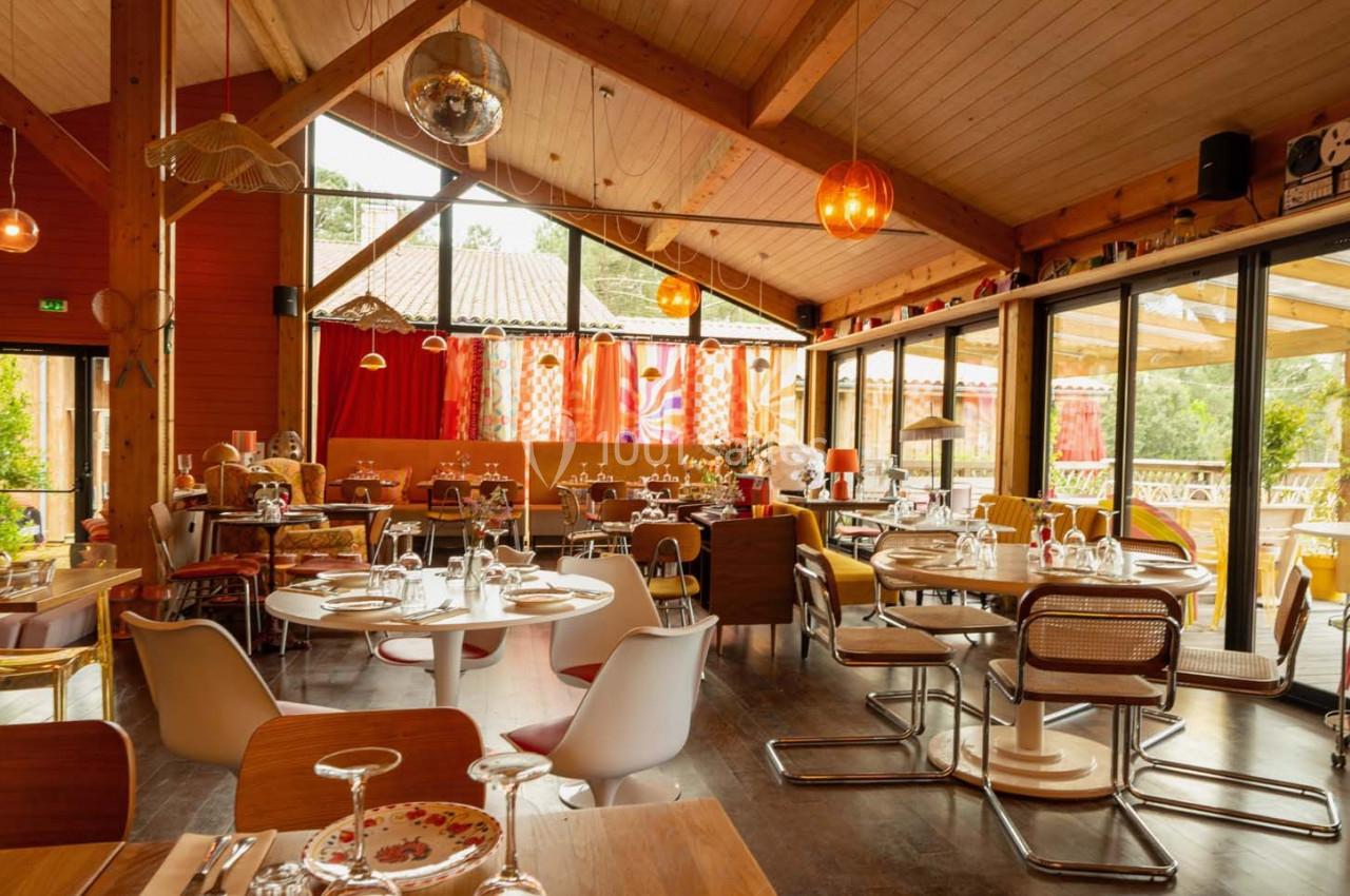 Salle de restaurant lumineuse avec tables dressées, grandes baies vitrées et décoration chaleureuse en bois.