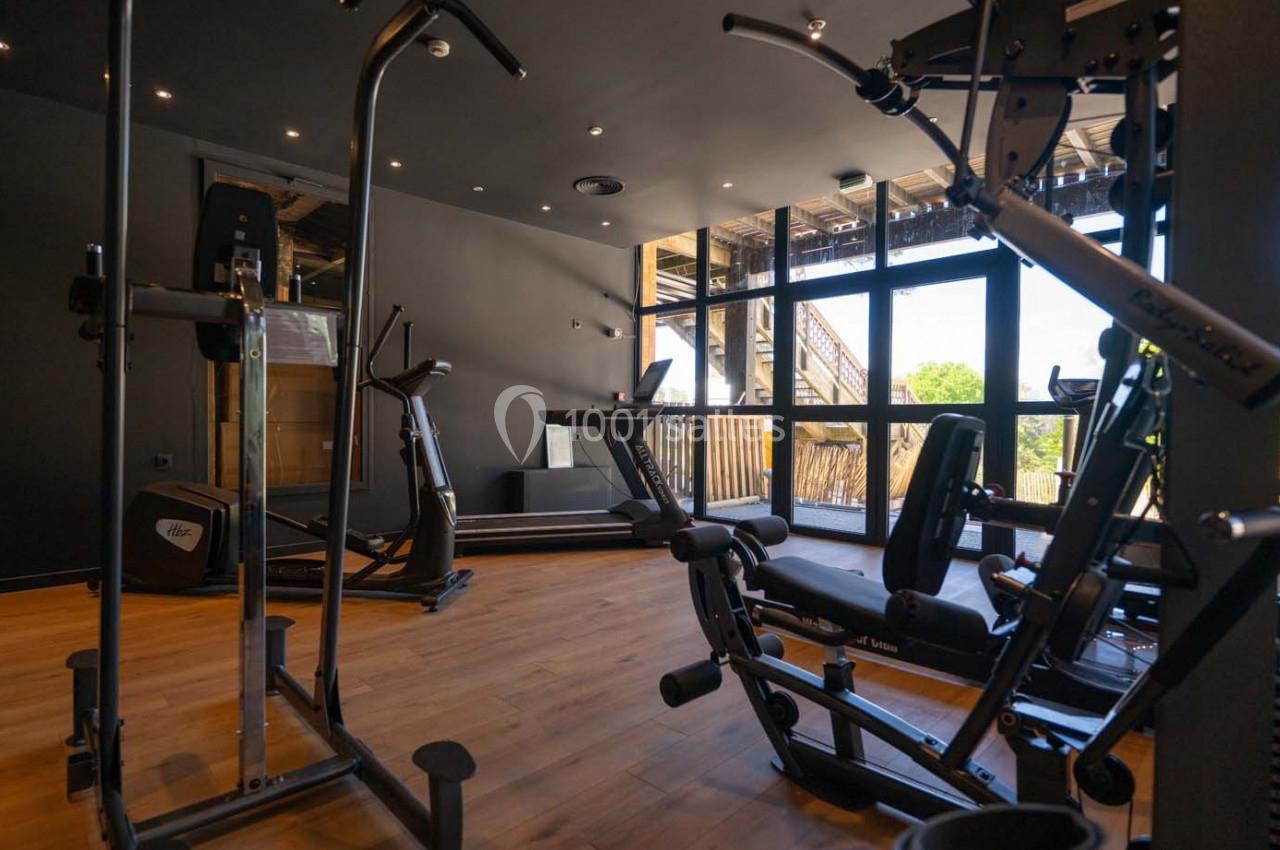 Salle de sport équipée de machines de musculation et de cardio, avec parquet et grandes fenêtres laissant entrer la lumière…