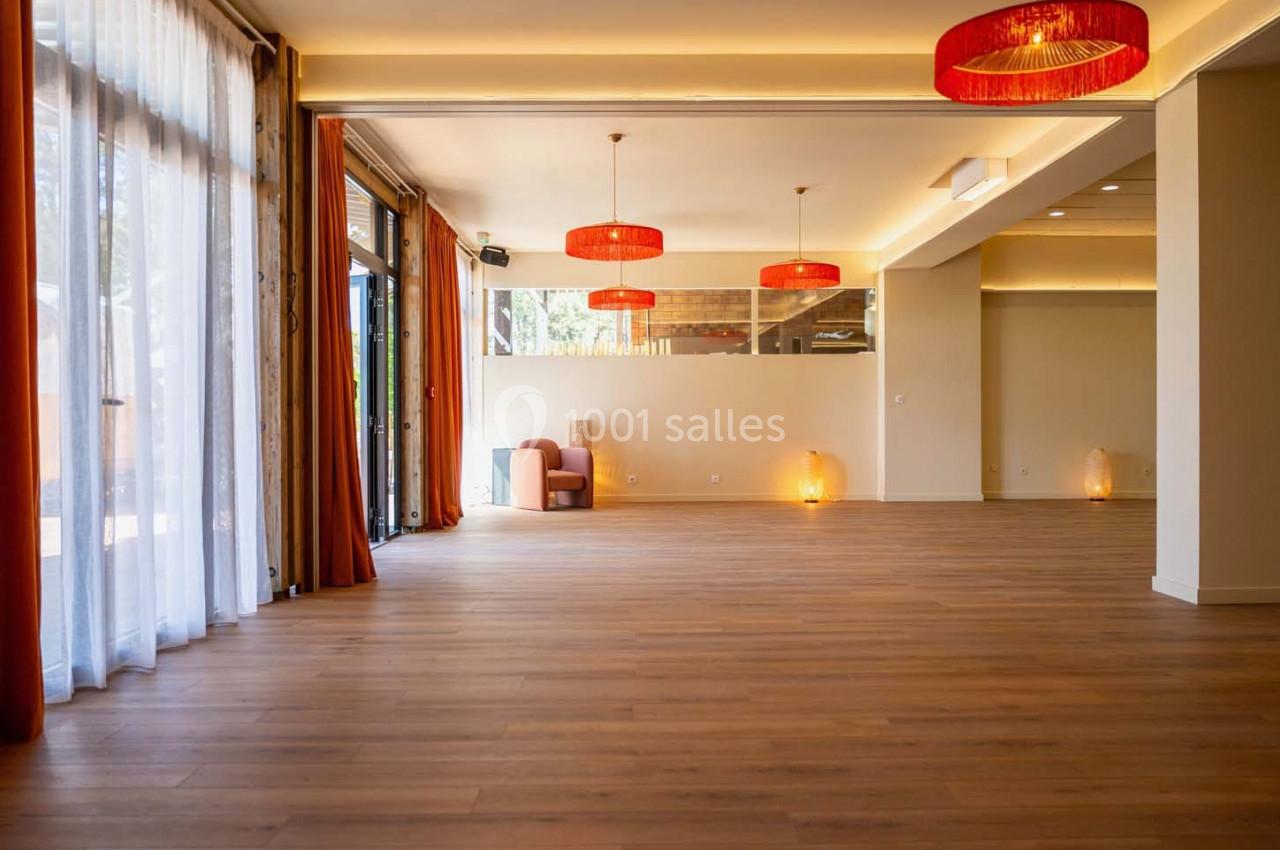Salle lumineuse avec parquet en bois, grandes baies vitrées, rideaux orange et suspensions rouges au plafond.