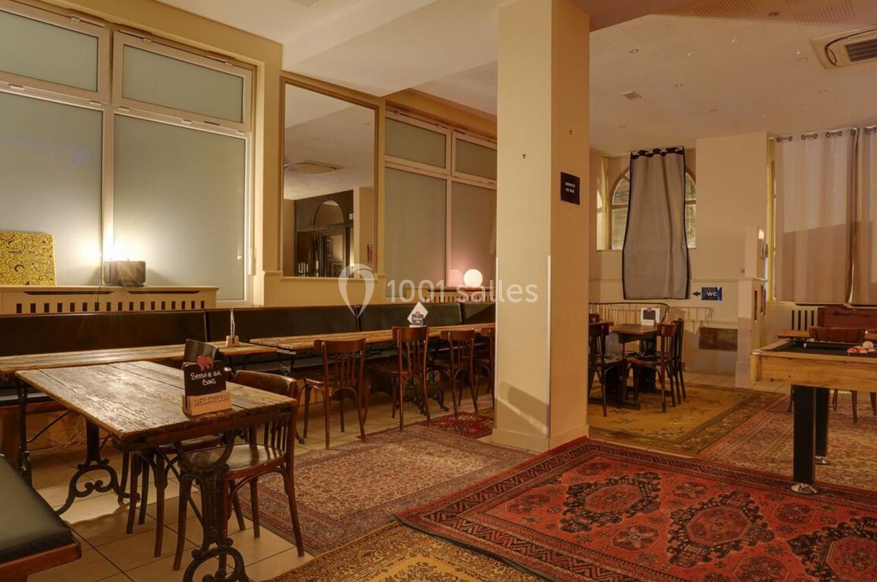 Salle avec tables en bois, chaises, banquettes, tapis colorés au sol et baby-foot dans un espace lumineux.