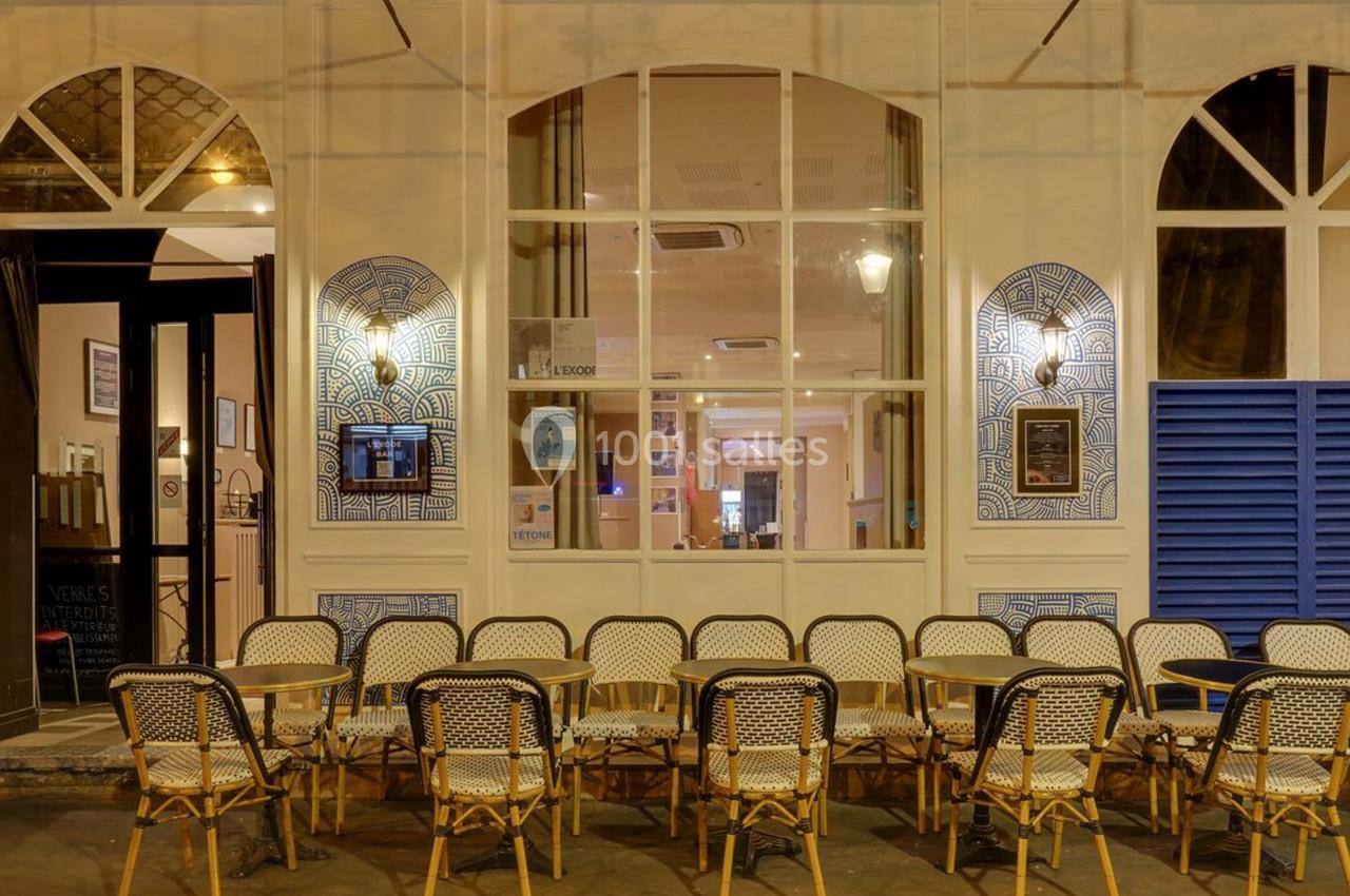 Façade d'un café avec des tables et chaises en terrasse, éclairée par des lampes murales en soirée.
