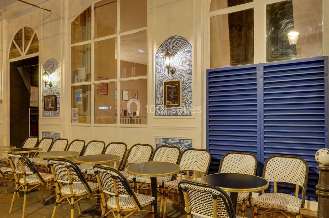 Terrasse extérieure d'un café avec des chaises en osier et des tables rondes, devant une façade éclairée le soir.