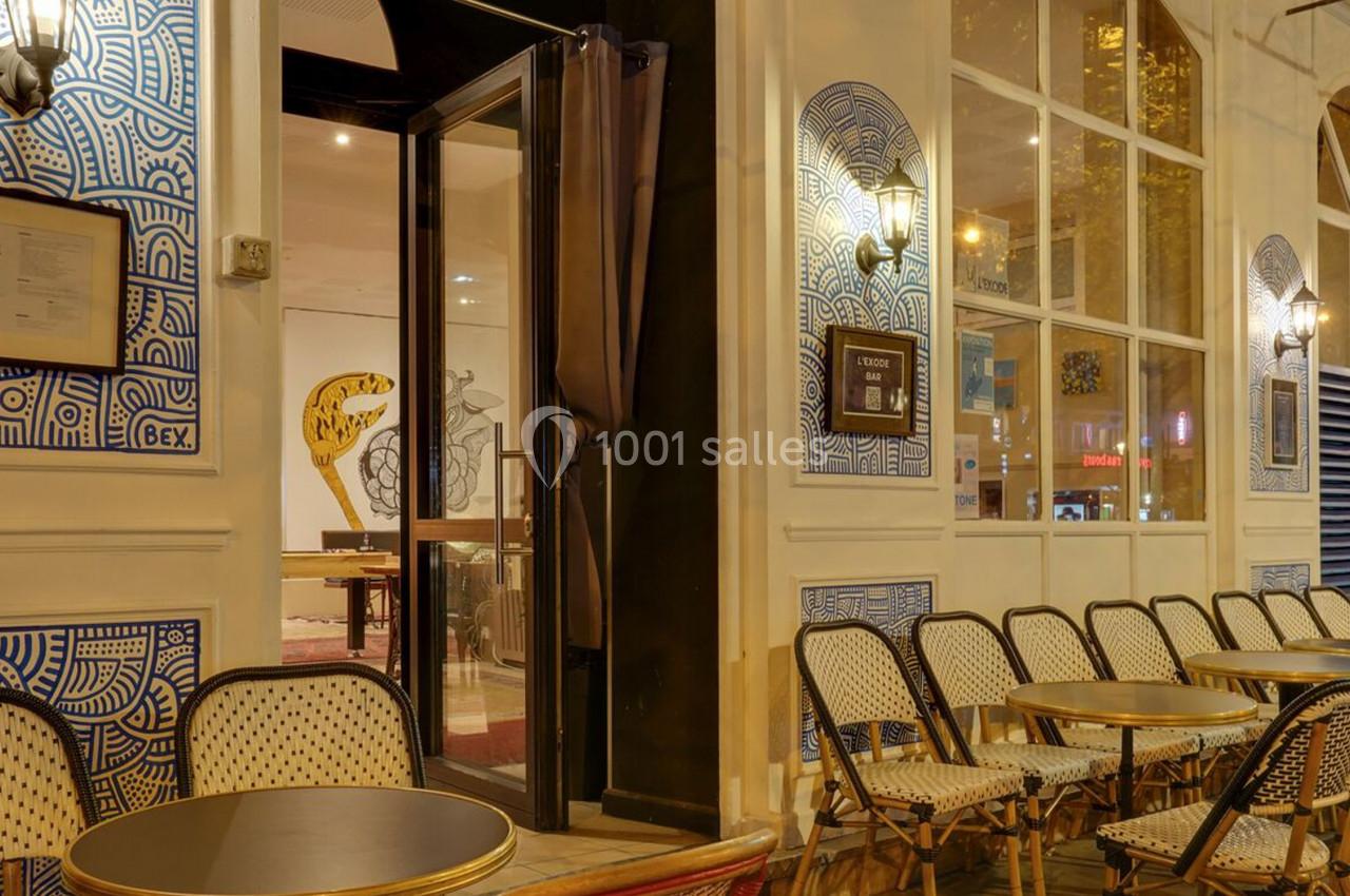Façade d'un café avec terrasse extérieure, chaises en osier et tables rondes alignées, éclairage chaleureux en soirée.