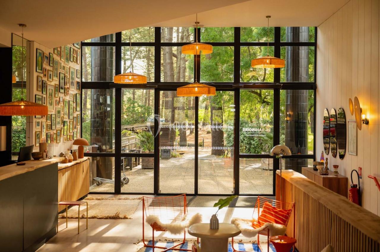 Hall lumineux avec de grandes baies vitrées, mobilier en bois et rotin, et vue sur un espace extérieur arboré.