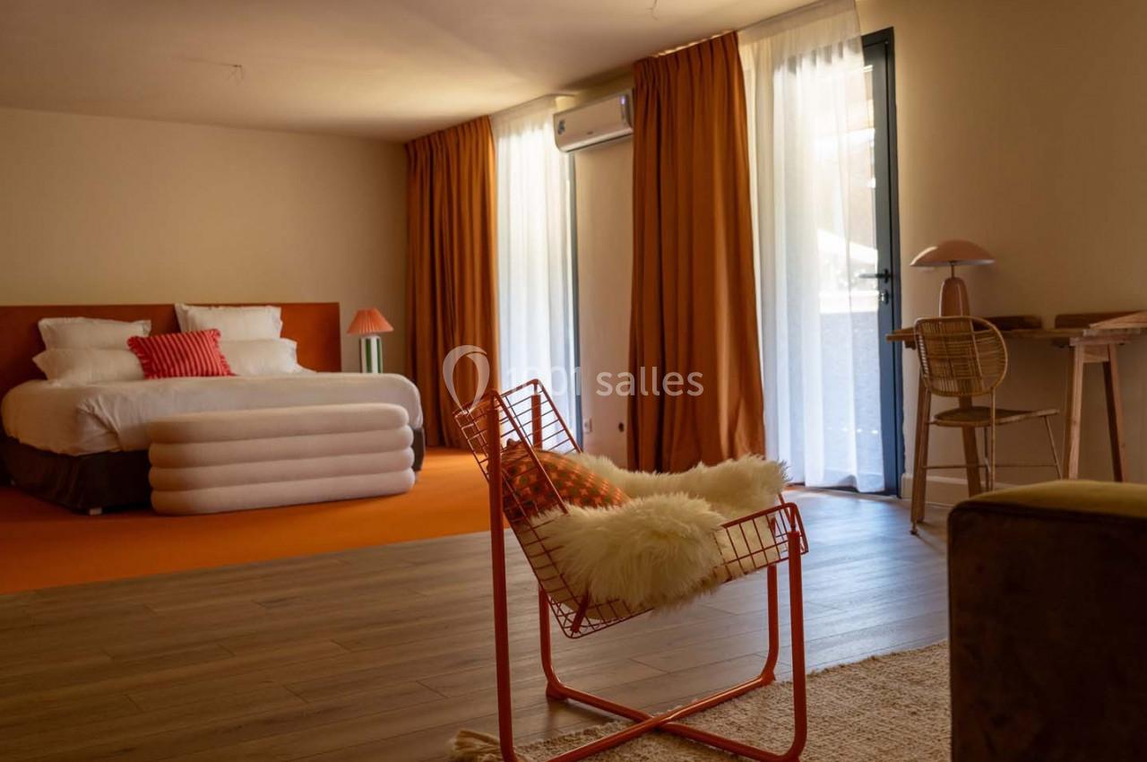 Chambre lumineuse avec lit double, fauteuil en métal, bureau en bois et rideaux orange devant une baie vitrée.