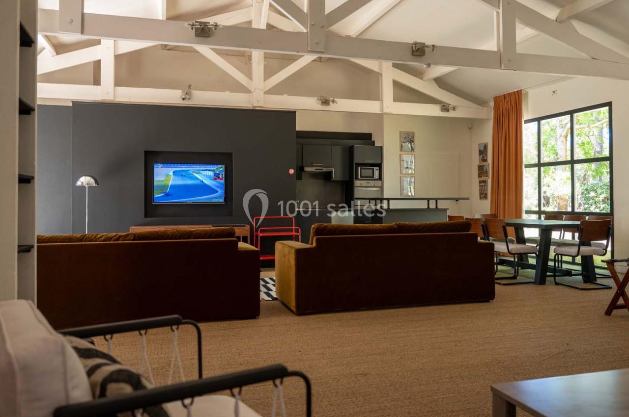 Salon spacieux avec canapés bruns, télévision allumée, cuisine ouverte et grande fenêtre laissant entrer la lumière…