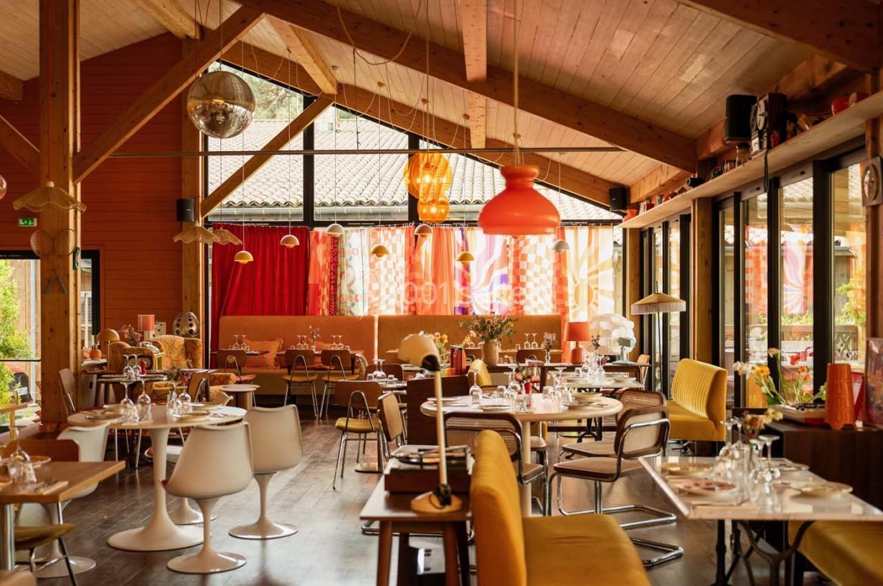 Salle de restaurant lumineuse avec décoration colorée, mobilier varié et grandes baies vitrées donnant sur l'extérieur.