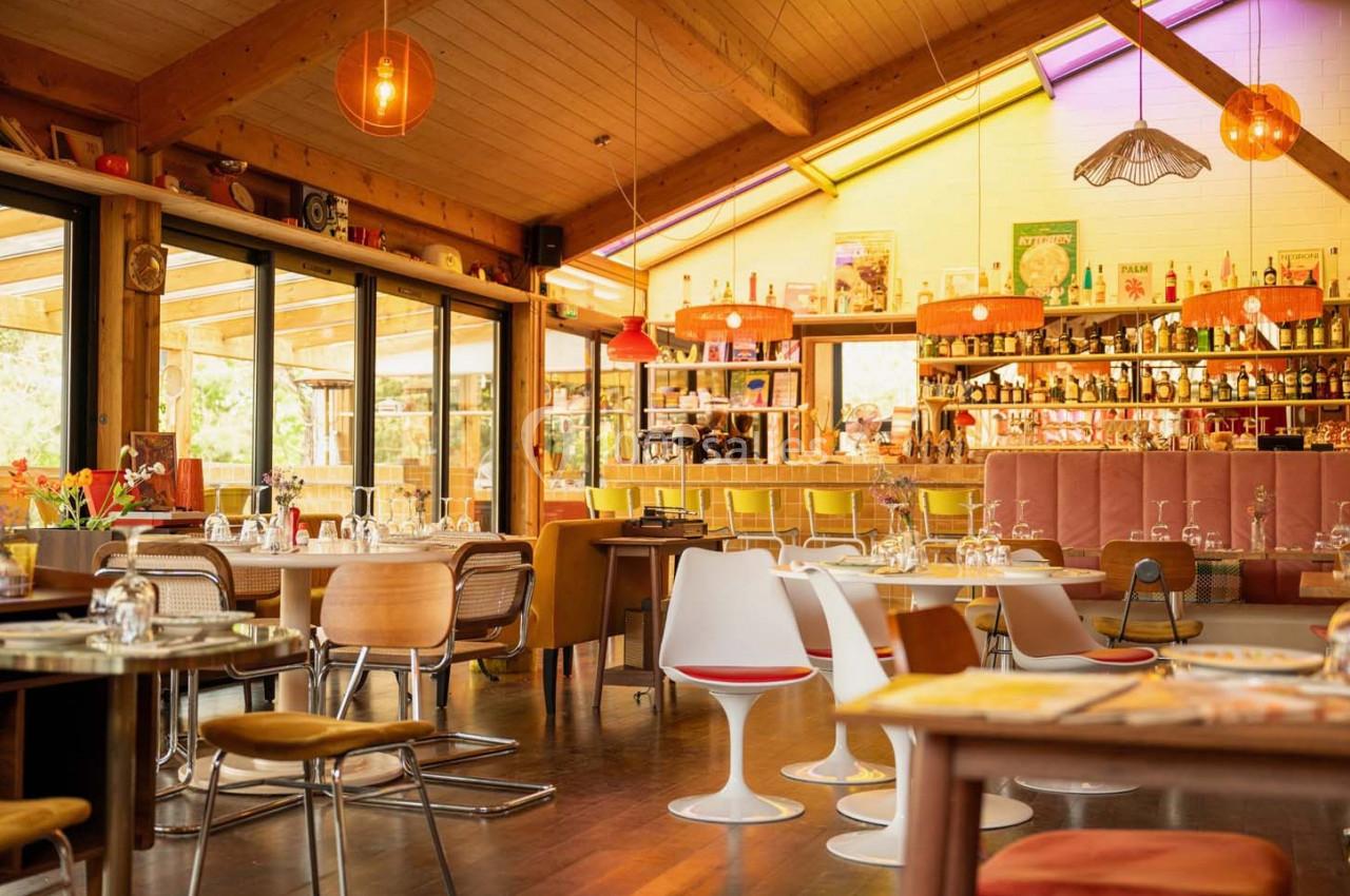 Salle de restaurant lumineuse avec mobilier coloré, grandes baies vitrées et bar en arrière-plan.