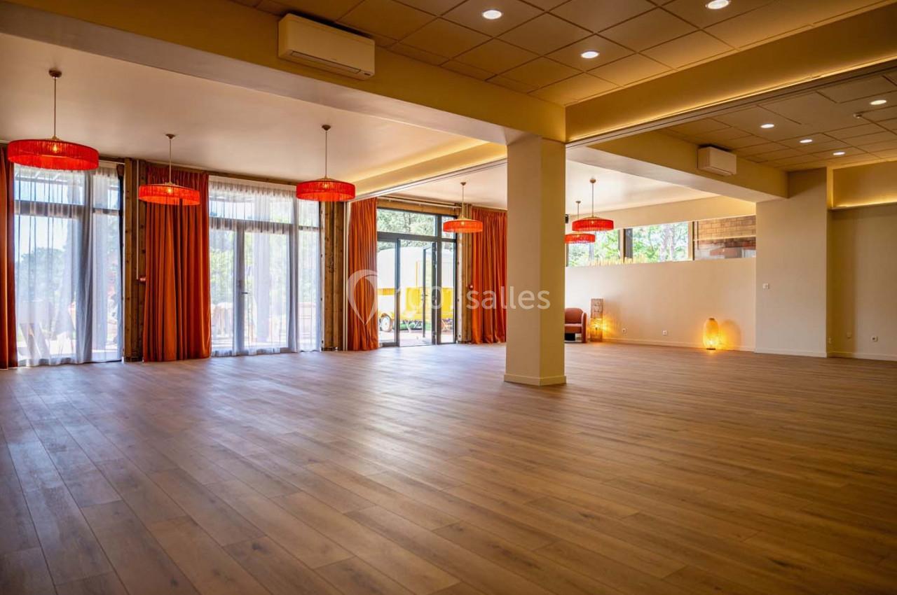 Salle spacieuse avec parquet en bois, grandes baies vitrées, rideaux orange et luminaires suspendus rouges.