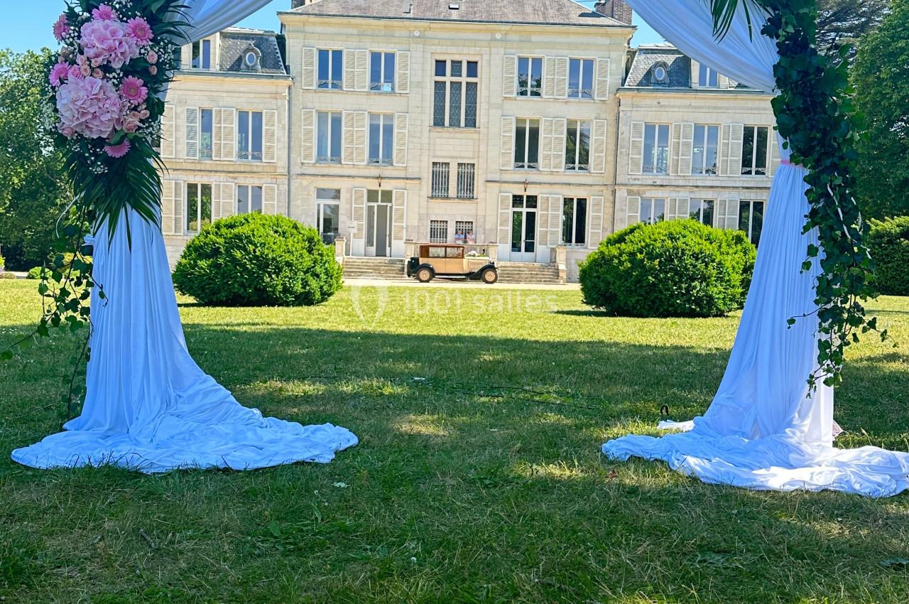 Arche de mariage décorée de fleurs et de drapés blancs, installée devant un château sous un ciel bleu.
