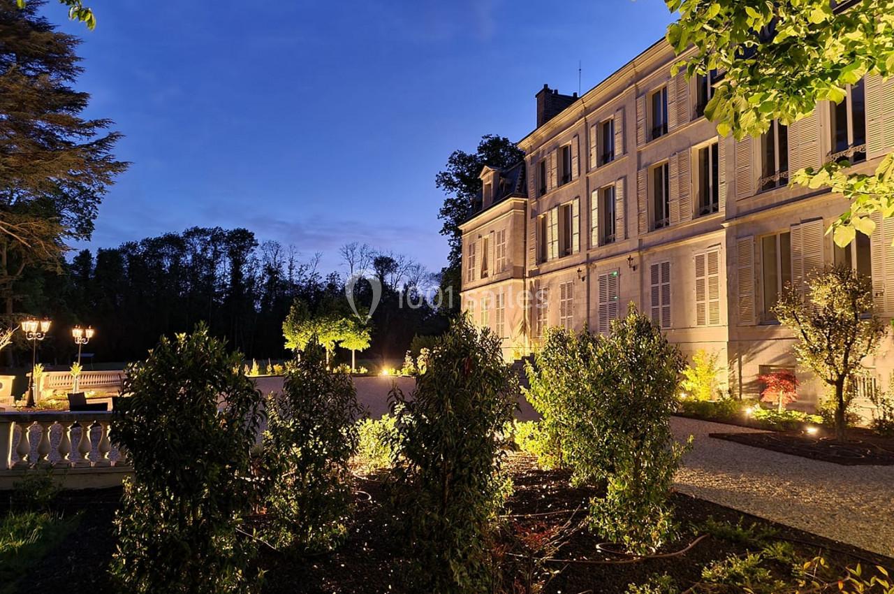 Façade d'un bâtiment historique éclairé au crépuscule, entouré de végétation et d'une allée gravillonnée.