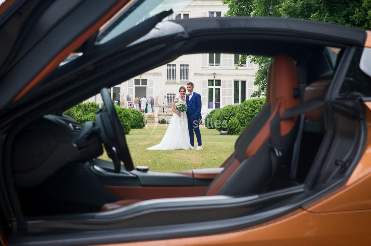 Vue d'un couple de mariés posant devant un bâtiment, cadrée à travers l'intérieur d'une voiture orange.
