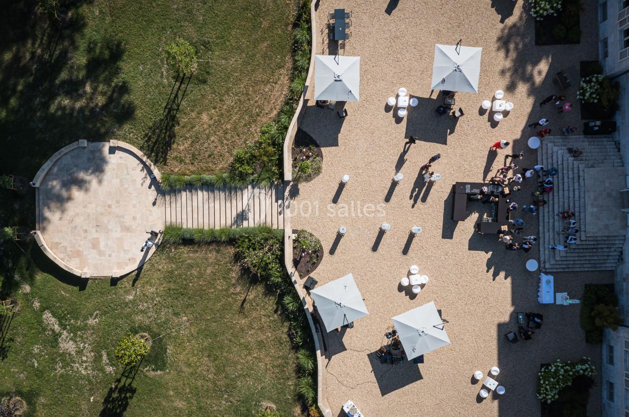 Vue aérienne d'une terrasse avec des parasols blancs, des tables et des personnes, entourée de pelouse et d'escaliers.