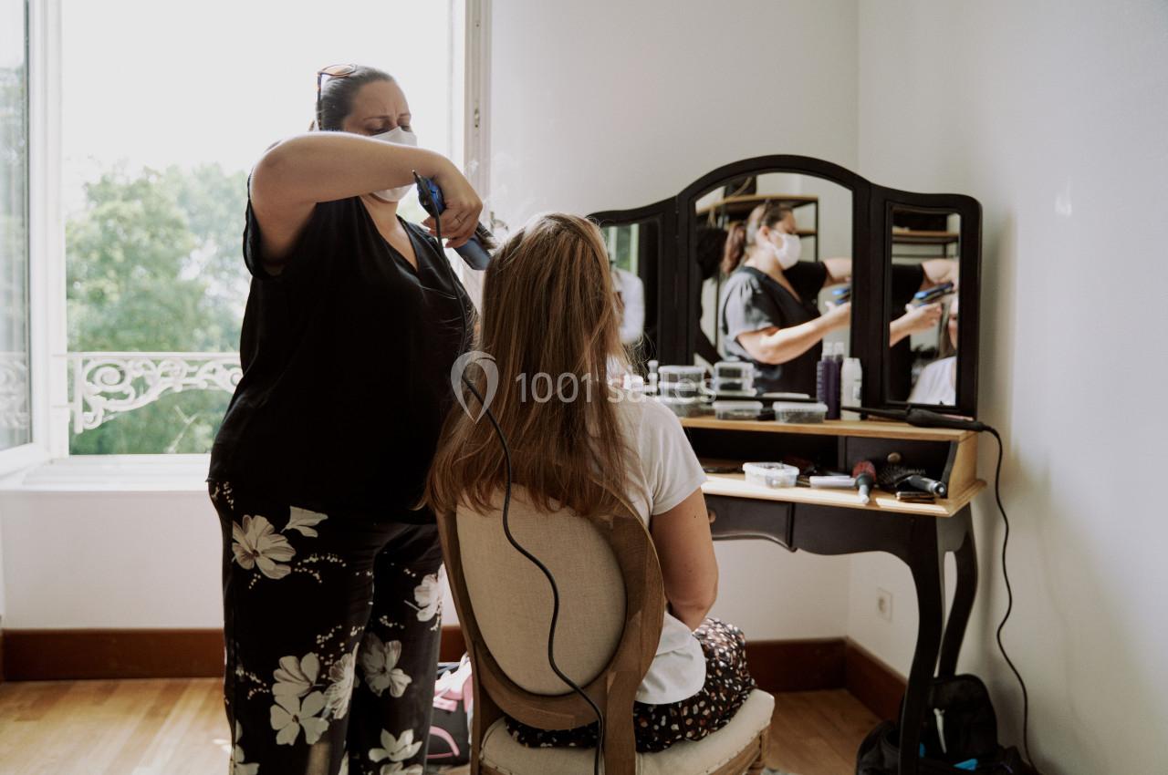 Une coiffeuse stylise les cheveux d'une femme assise devant une coiffeuse dans une pièce lumineuse.