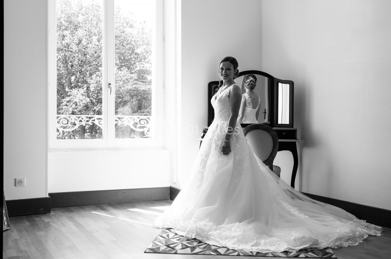 Femme en robe de mariée souriant devant un miroir dans une pièce lumineuse avec une grande fenêtre.