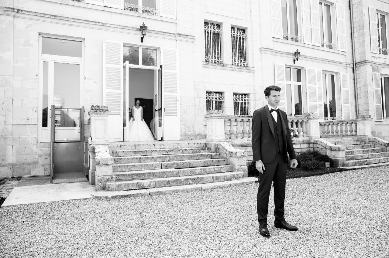 Un homme en costume se tient dans une cour pavée devant un bâtiment, une femme en robe de mariée sort par une porte.