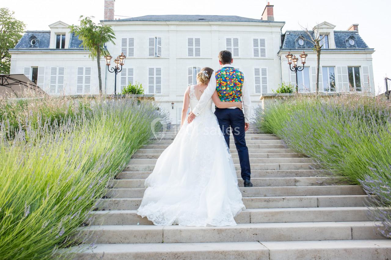 Un couple en tenue de mariage monte un escalier bordé de lavande, avec un grand bâtiment blanc en arrière-plan.