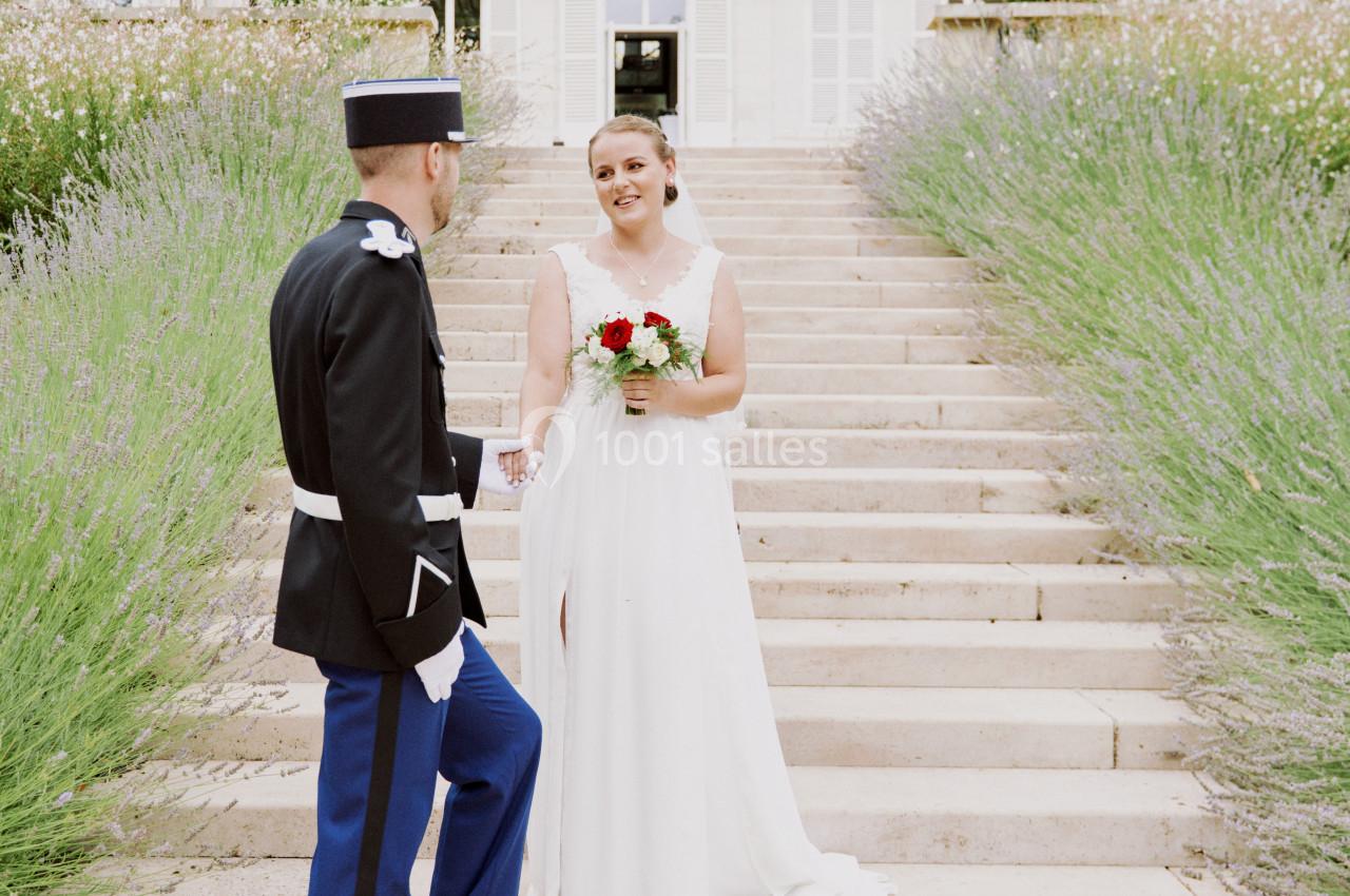 Une mariée en robe blanche tient un bouquet face à un homme en uniforme devant un escalier bordé de lavande.