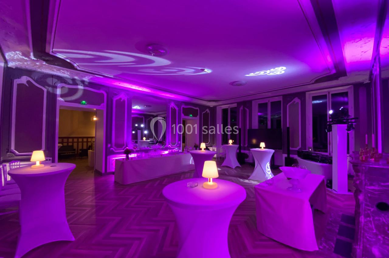 Salle de réception décorée avec des tables hautes, éclairage violet et petites lampes allumées.
