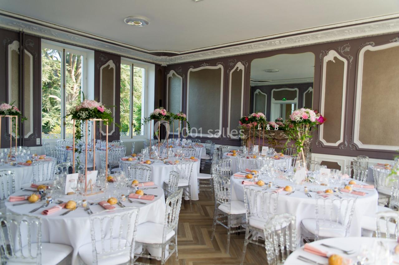 Salle de réception élégante avec tables rondes dressées, décor floral et chaises transparentes.
