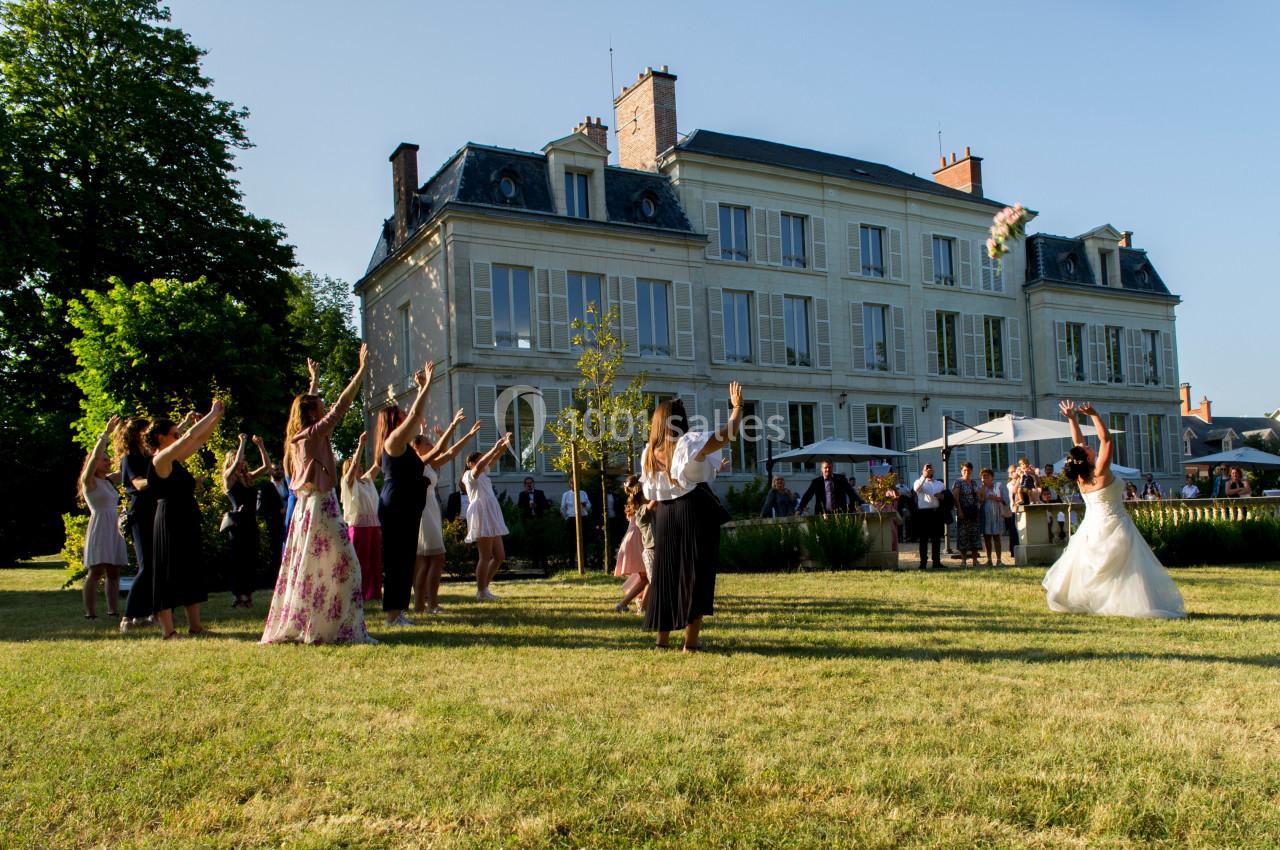 Une mariée lance son bouquet à un groupe d'invitées dans le jardin d'un grand bâtiment élégant par temps ensoleillé.