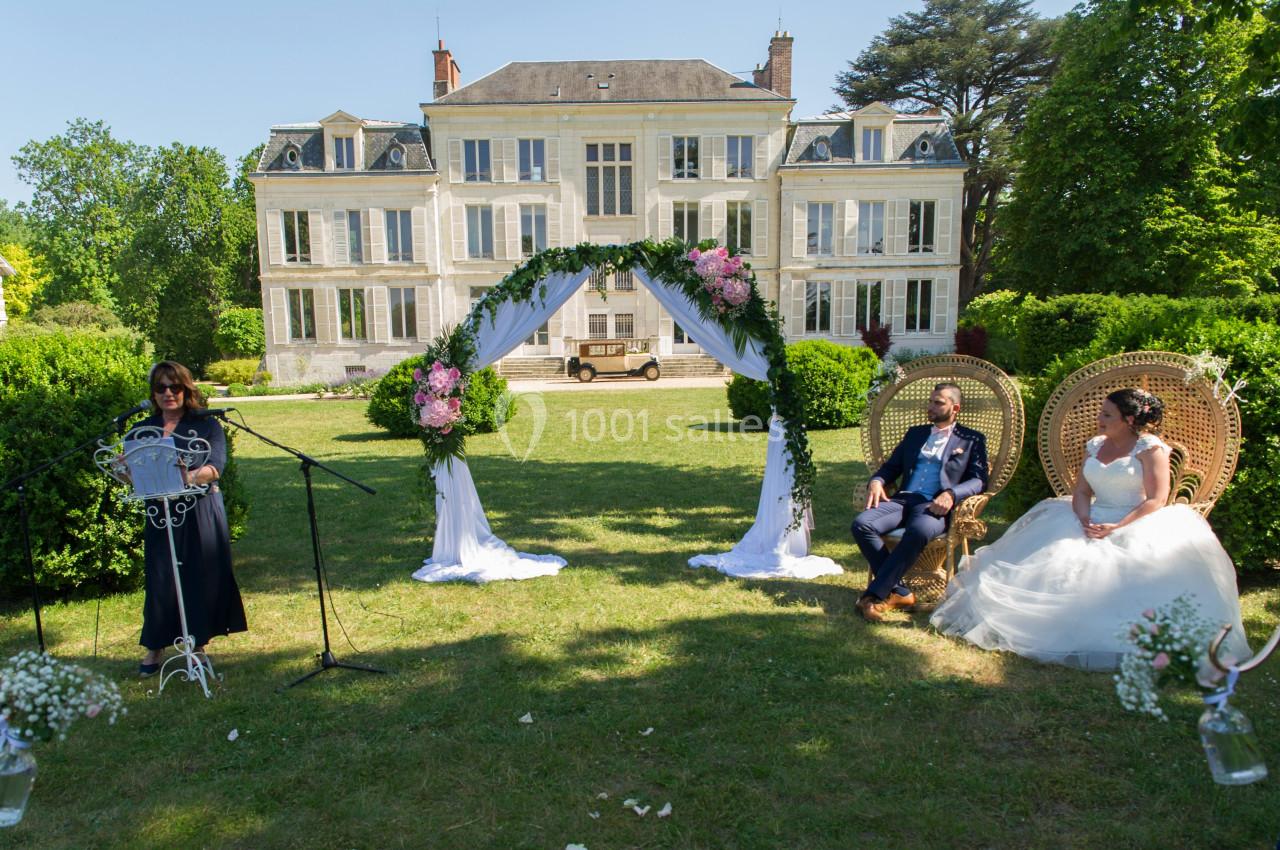 Cérémonie de mariage en extérieur devant un manoir, avec une officiant, un couple assis et une arche décorée de fleurs.
