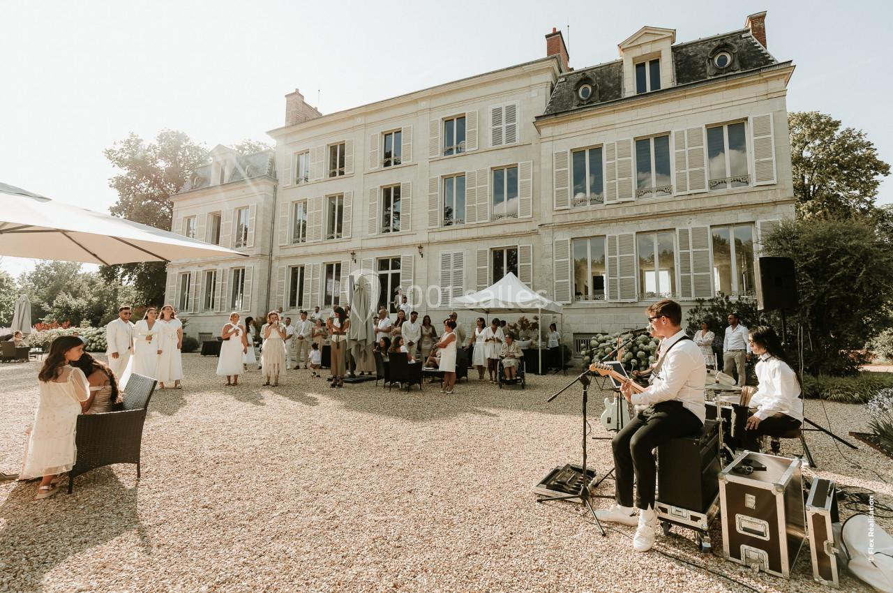 Un groupe de musiciens joue en plein air devant un grand bâtiment de style classique, entouré d'invités en tenue claire.