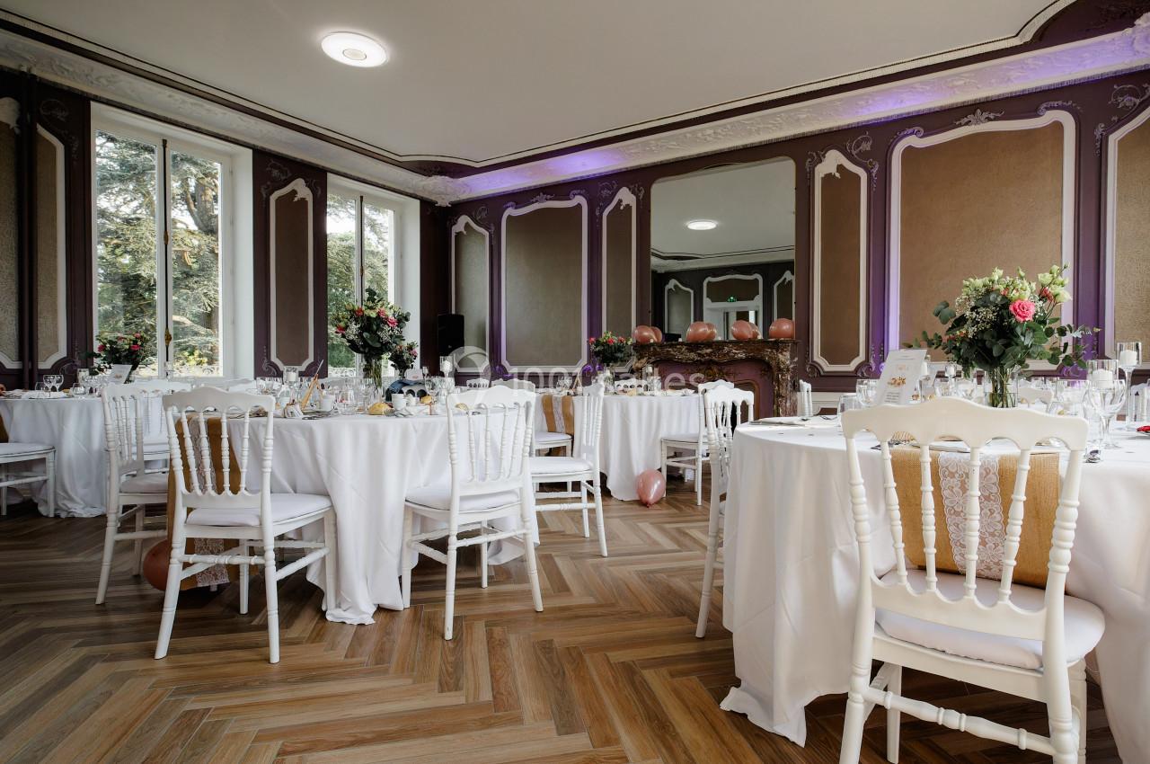 Salle de réception élégante avec tables rondes dressées, chaises blanches et décorations florales, parquet au sol.
