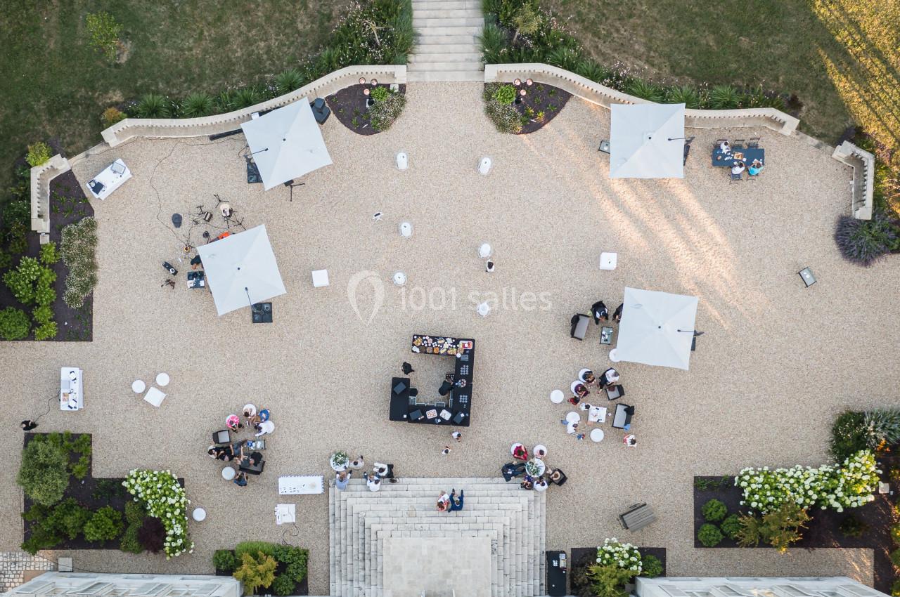 Vue aérienne d'une terrasse avec des tables, des parasols blancs et un bar central entouré de personnes.