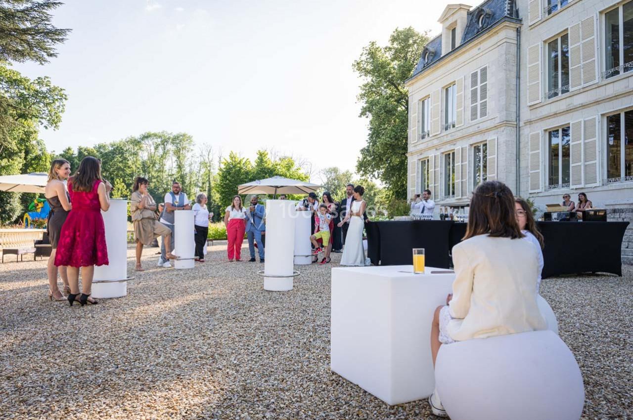 Personnes rassemblées dans une cour gravillonnée devant un bâtiment élégant, discutant autour de tables hautes.