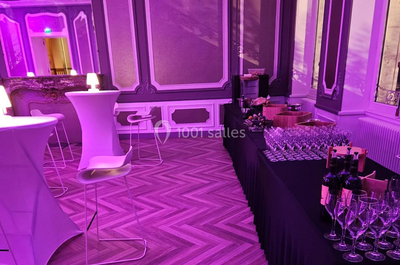 Salle élégante avec tables hautes, chaises modernes et buffet de verres et bouteilles de vin, éclairée par une lumière…