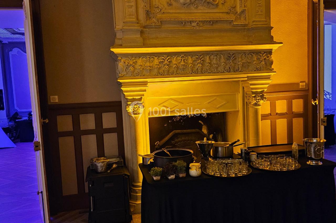 Table dressée avec verres, herbes et ustensiles devant une cheminée décorative dans une pièce éclairée.