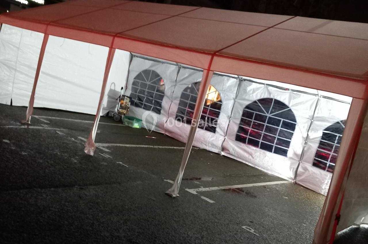 Tente blanche avec parois transparentes installée sur un parking, éclairée de l'intérieur en soirée.