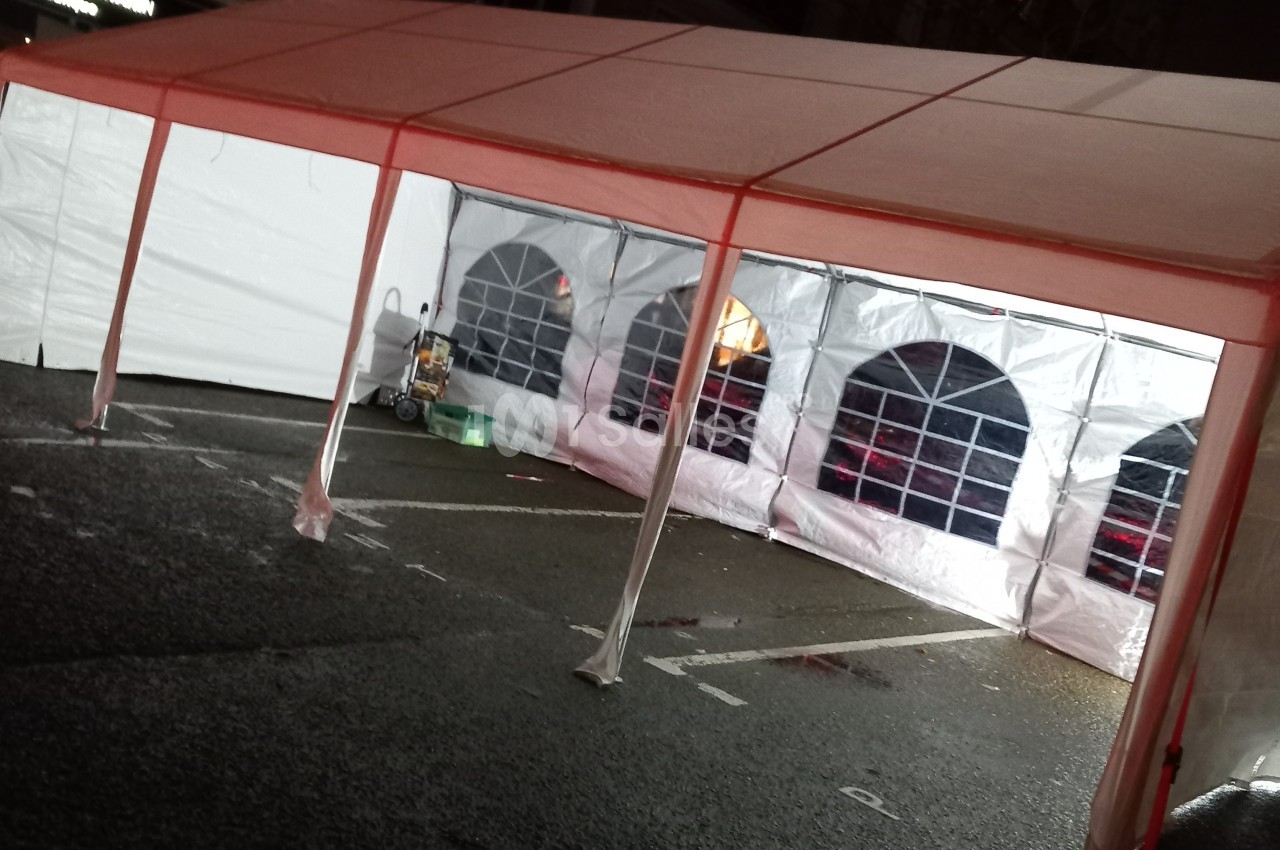Tente blanche avec parois transparentes installée sur un parking, éclairée de l'intérieur en soirée.