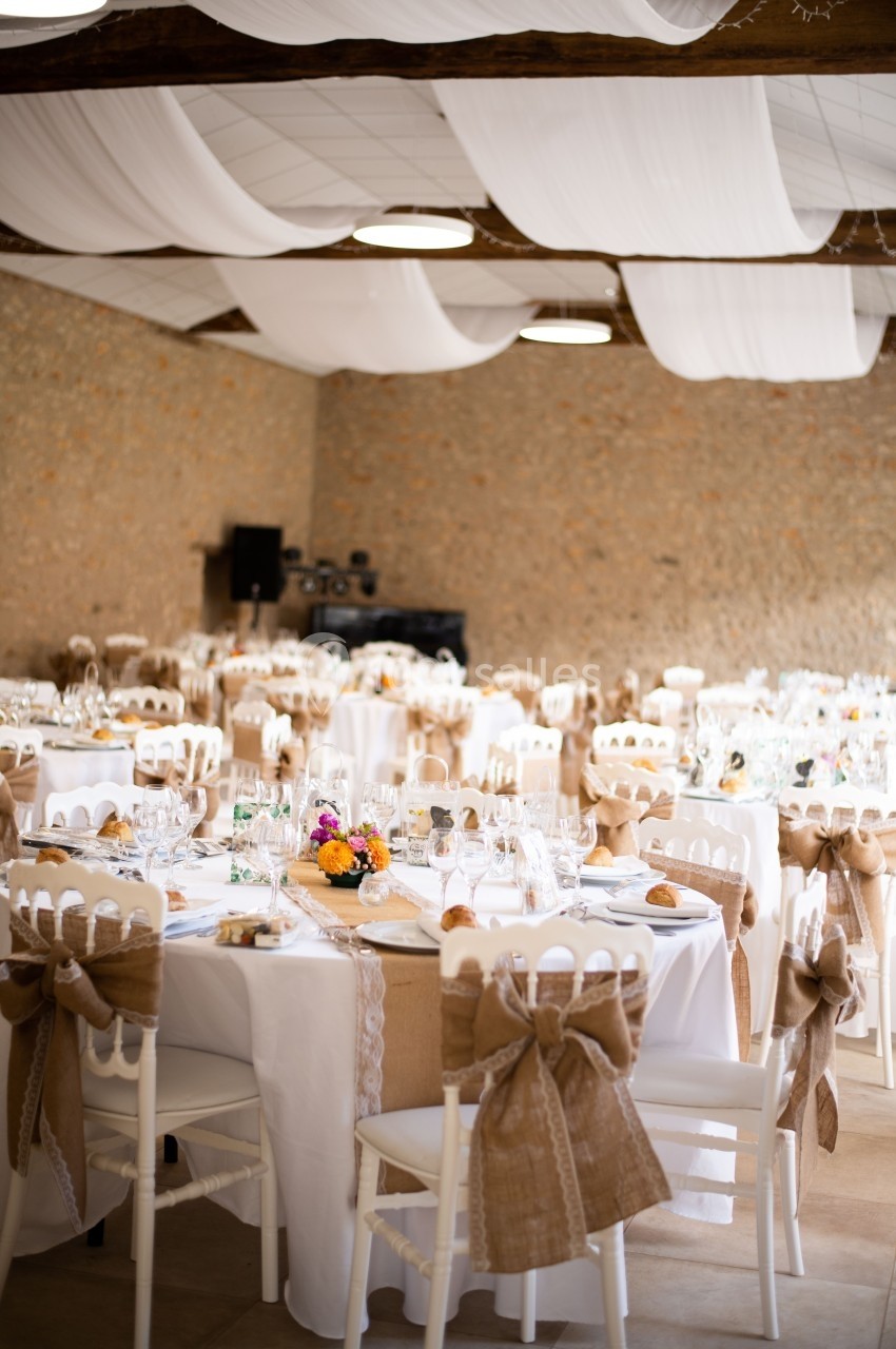 Salle de réception décorée pour un mariage, avec tables dressées, chaises ornées de nœuds en toile de jute et plafond drapé.