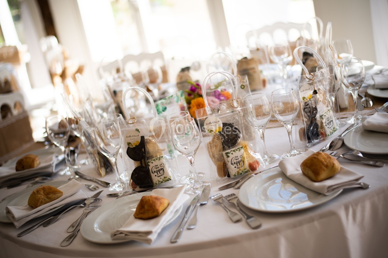 Table dressée pour un repas, avec assiettes, verres, couverts et sachets cadeaux disposés sur une nappe blanche.