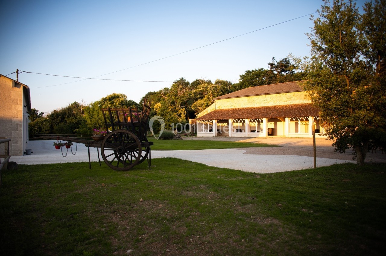 Cour extérieure avec une charrette ancienne en bois, pelouse et bâtiment en pierre éclairé par une lumière de fin de journée.