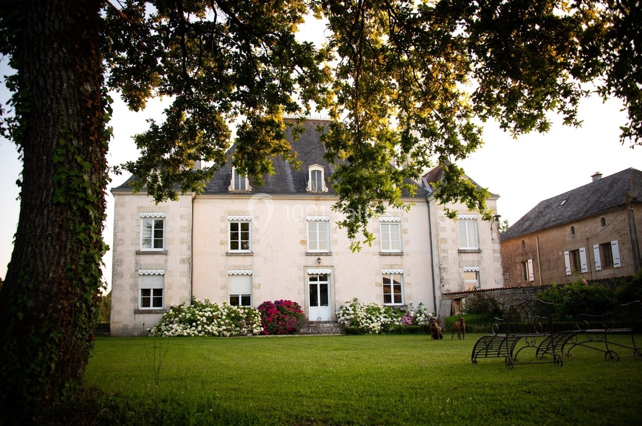 Façade d'un manoir ancien entouré d'un jardin verdoyant avec des fleurs et des arbres en premier plan.