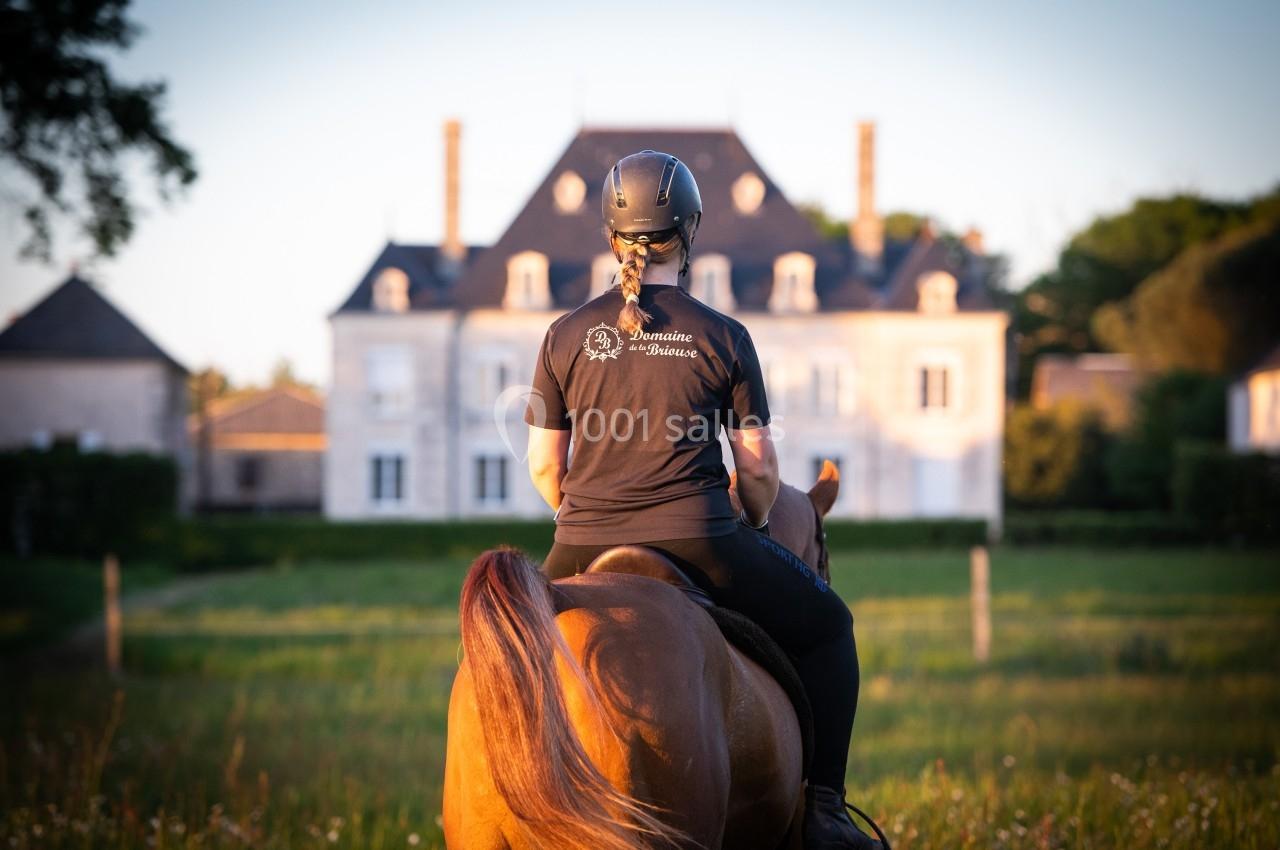 Une cavalière vue de dos, montant un cheval dans un pré, avec un manoir en arrière-plan au coucher du soleil.