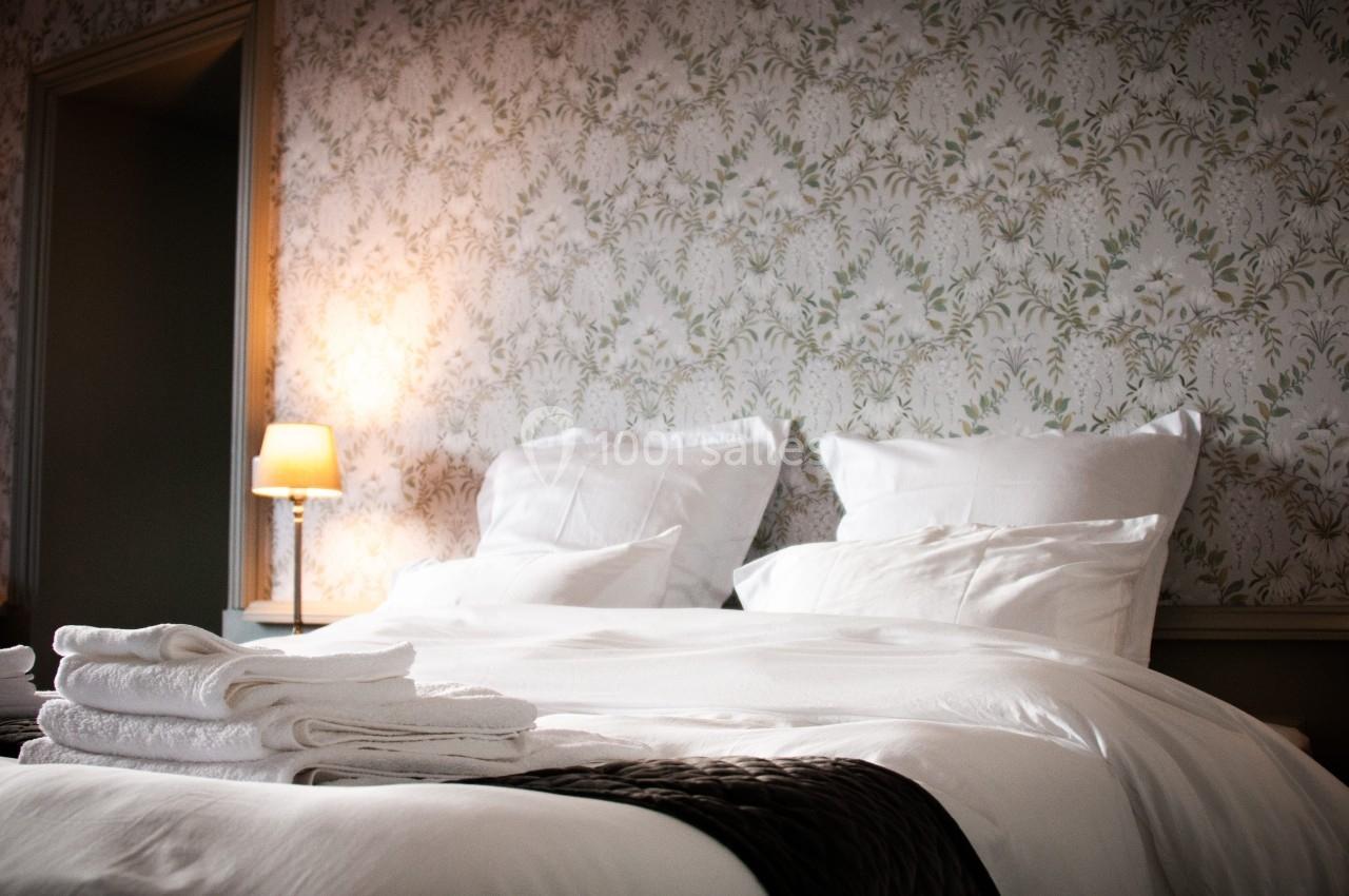 Lit double avec draps blancs, oreillers moelleux, serviettes pliées au premier plan et lampe allumée en arrière-plan.